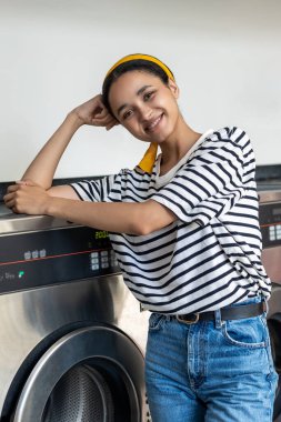 Çamaşır odasında poz veren genç esmer kadın çamaşır makinesine yaslanıyor.