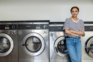 Çamaşır odasında poz veren genç esmer kadın çamaşır makinesine yaslanıyor.