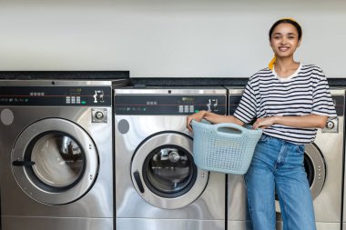 Çamaşırhanede çamaşır makinesini kullanan gülümseyen bir kadın temizlik için kirli çamaşırları yıkıyor.