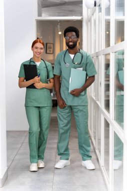 Kızıl saçlı kadın sıhhiyeci, hastane koridorunda Afro-Amerikan erkek meslektaşıyla duruyor.