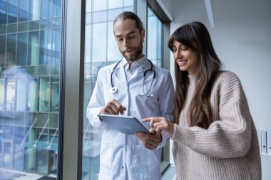 Klinik koridorunda kadın hastayla dijital danışma için elinde tablet bulunan erkek doktor.