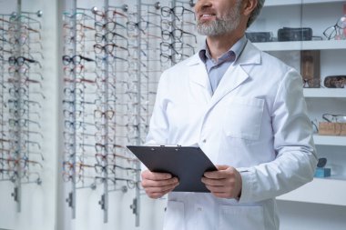 Optik. Beyaz laboratuvar önlüklü bir adam gözlükçünün yanında dikiliyor.