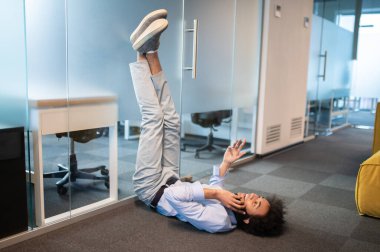 Rahatlamış genç adam, bacaklarını kaldırarak yerde uzanıyor ve ofiste cep telefonu kullanıyor.