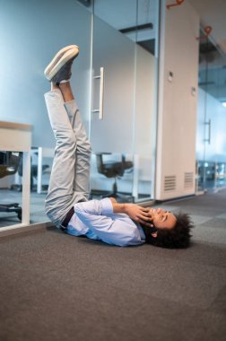 Rahatlamış genç bir adam, bacaklarını kaldırarak yerde uzanıyor ve ofiste cep telefonuyla konuşuyor.