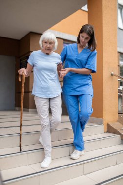 Yaşlı hastaya bastonla yardım eden bir tıp çalışanı kıdemli hastaya yardım öneriyor.