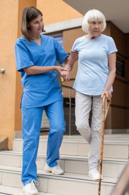 Yaşlı bir hastayla yürüyen kadın hemşire emekliliğe destek olmak için baston kullanıyor.