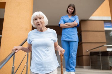 Yaşlı hastaya bastonla yardım eden bir tıp çalışanı kıdemli hastaya yardım öneriyor.