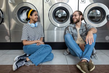 Çamaşırhanedeki çift otomatik çamaşır makinesinin çamaşır işlerini bitirmesini beklerken konuşuyor.