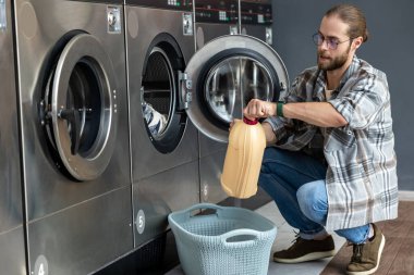 Ticari çamaşırhanedeki sakallı adam ev kıyafetlerini temizlemek için otomatik çamaşır makinesindeki deterjanı kullanıyor.