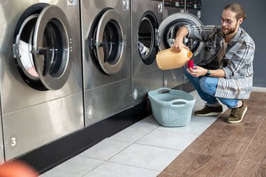 Ticari çamaşırhanedeki sakallı adam ev kıyafetlerini temizlemek için otomatik çamaşır makinesindeki deterjanı kullanıyor.