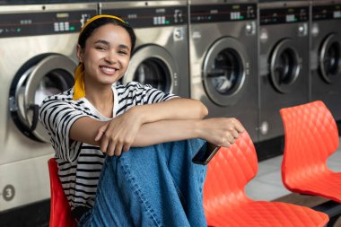Çamaşırhanede sıralanmış gülümseyen bir kadın çamaşır makinesinin yanında oturmuş çamaşırların yıkanmasını bekliyor.