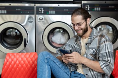 Çamaşırhanede gülümseyen sakallı bir adam cep telefonunda internette dolaşırken çamaşır makinesi kullanıyor.