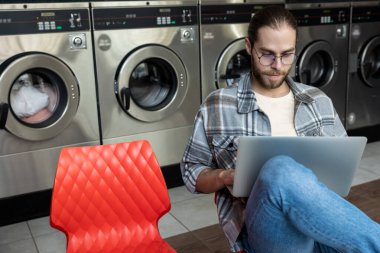 Çamaşırhanedeki adam çamaşır makinelerinin yanında otururken çamaşırların yıkanmasını ve dizüstü bilgisayarın çevrimiçi işlerde kullanılmasını bekliyor.