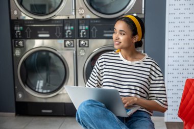 Çamaşırhanede çamaşır makinesinin yanında oturan bir kadın çevrimiçi iş üzerinde çalışırken dizüstü bilgisayar kullanıyor.