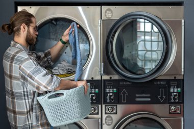 Çamaşırhanedeki sakallı adam çamaşırlarını yıkarken ticari çamaşır makinesi kullanıyor.