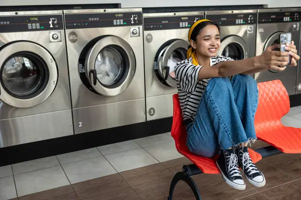 Çamaşırhanede gülümseyen ev hanımı çamaşır makinesi kullanırken cep telefonundan selfie çekiyor.