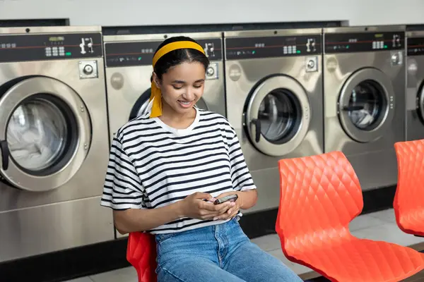 Çamaşırhanede gülümseyen ev hanımı cep telefonunda gezinirken çamaşır makinesi kullanıyor.
