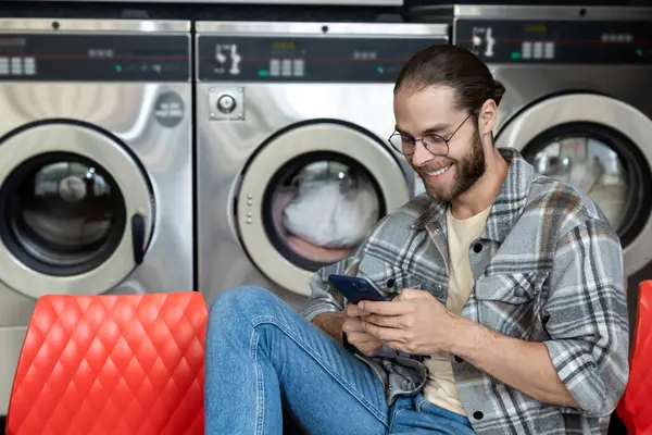 Çamaşırhanede gülümseyen sakallı bir adam cep telefonunda internette dolaşırken çamaşır makinesi kullanıyor.
