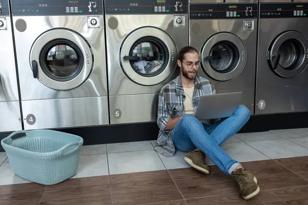 Çamaşırhanede çamaşır makinesinin yanında oturan bir adam çevrimiçi iş üzerinde çalışırken dizüstü bilgisayar kullanıyor.