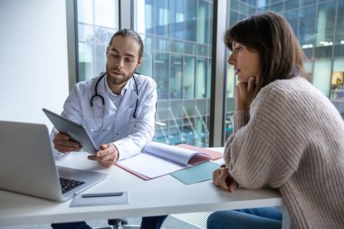 Klinikteki sakallı doktor kadın hastayla görüşürken elinde tablet tutuyor.