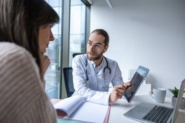 Klinikteki sakallı doktor kadın hastayla görüşürken elinde tablet tutuyor.