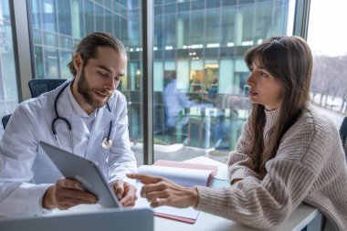 Laboratuvardaki erkek doktor tıbbi danışmanlık için dokunmatik ekran tableti kullanıyor. Kadın hastalarla test sonuçlarını analiz ediyor.