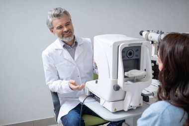Gri saçlı erkek doktor klinikte profesyonel teknolojiyle kadın hasta gözlerini muayene ediyor.