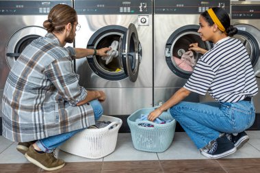 Erkek ve kadın birlikte ticari çamaşırhanede otomatik çamaşır makinesi ve kurutucuyu ev kıyafetlerini temizlemek için kullanıyorlar.