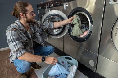 Kamu çamaşırhanesinde modern teknolojiyi kullanan sakallı bir adam çamaşır makinesinde çamaşır yıkıyor.