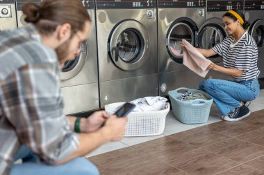Çamaşırhanedeki bir çift, çamaşır yıkarken self-servis teknolojisi kullanan bir adamla sohbet ediyor ve cep telefonunu kontrol ediyor.