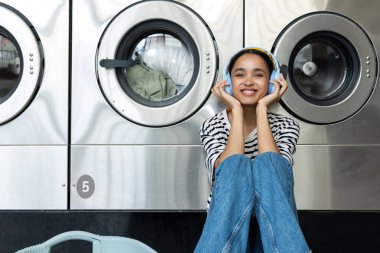 Genç bir kadın otomatik çamaşırhanede oturmuş kulaklıkla müzik dinliyor.