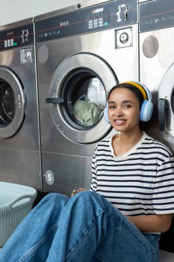 Genç bir kadın otomatik çamaşırhanede oturmuş kulaklıkla müzik dinliyor.