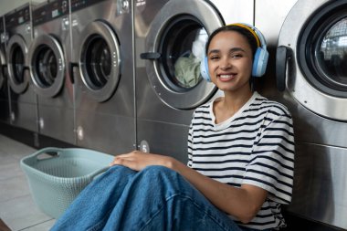 Çamaşırhanede oturmuş kulaklık takmış gülümseyen bir kadın çamaşır makinesinin temizliğini bitirmesini bekliyor.
