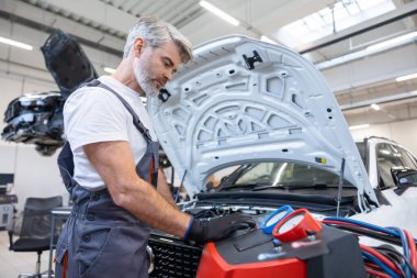 Servis atölyesindeki sakallı tamirci, arabanın klima sistemindeki ısı seviyesini ölçen cihazı kullanıyor.
