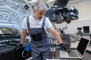 Sakallı teknisyen, arabanın kaputunun altındaki teftiş aracının detaylarına bakıyor.