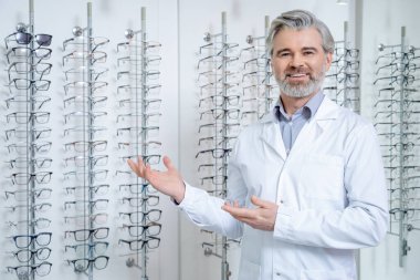 Optik gemi. Gri saçlı, yetişkin bir erkek göz doktoru. Standın yanında gözlüklü duruyor.
