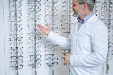 Optik gemi. Gri saçlı, yetişkin bir erkek göz doktoru. Standın yanında gözlüklü duruyor.