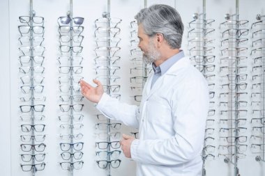 Optik gemi. Gri saçlı, yetişkin bir erkek göz doktoru. Standın yanında gözlüklü duruyor.