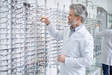 Oftalmoloji. Laboratuvar önlüklü beyaz erkek göz doktoru standın yanında gözlüklü duruyor.