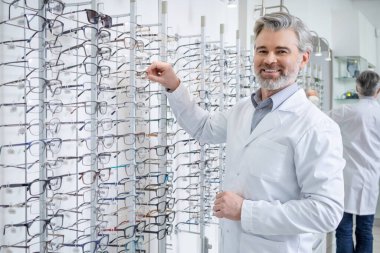 Oftalmoloji. Laboratuvar önlüklü beyaz erkek göz doktoru standın yanında gözlüklü duruyor.