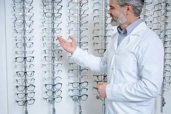 Optik gemi. Gri saçlı, yetişkin bir erkek göz doktoru. Standın yanında gözlüklü duruyor.