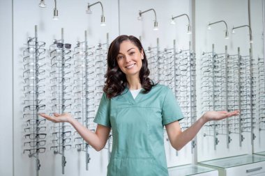 Optik dükkanında. Genç bayan göz doktoru gözlük standının yanında duruyor ve yeni modeller gösteriyor.
