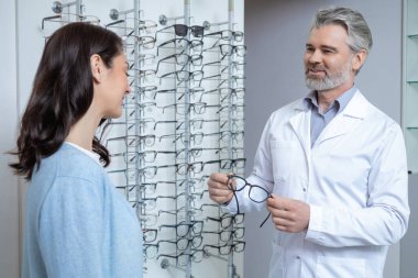 Esmer kadın optik mağazada gözlük seçiyor ve göz doktoruyla görüşünü kontrol ediyor.