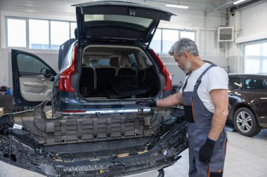 Gri saçlı, üniformalı erkek tamirci garajda araç kullanıyor. Tamir için otomobili kontrol ediyor.