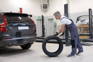 Araba tamirhanesinde yeni lastik tutan beyaz üniformalı erkek tamirci tamir işi yapıyor.