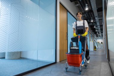 Temizlik personeli. Siyah saçlı, üniformalı, temizlik malzemeleriyle dolu bir arabası olan genç bir kadın.