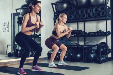 Çömelme. Spor yaparken spor salonunda çömelen iki genç kadın.