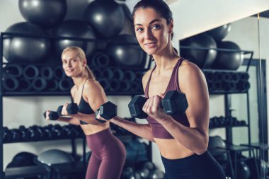 Dambıllarla egzersiz. İki genç beyaz kadın dambıllarıyla egzersiz yapıyor ve odaklanmış görünüyor.