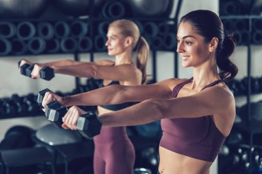 Dambıllarla egzersiz. İki genç beyaz kadın dambıllarıyla egzersiz yapıyor ve odaklanmış görünüyor.