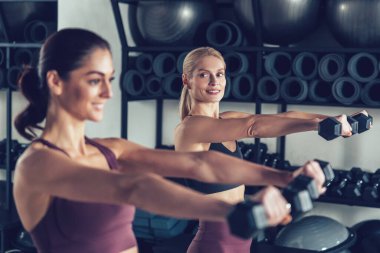 Dambıllarla egzersiz. İki genç beyaz kadın dambıllarıyla egzersiz yapıyor ve odaklanmış görünüyor.